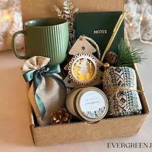 May include: A gift box filled with winter-themed items, including a green mug, a burlap bag with a green ribbon, a wooden Christmas tree ornament, a candle with a gold lid and a label that says "Winter Cranberry", and a pair of green and white patterned socks. The box is brown with the words "EVERGREEN JOY" printed on the bottom.