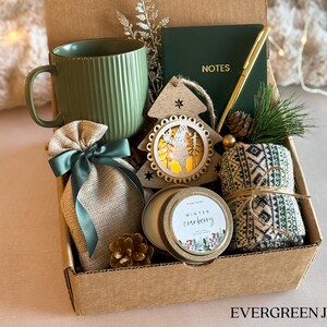 Puede incluir: Una caja de regalo llena con una taza verde, un cuaderno peque&ntilde;o con el texto "NOTES", un bol&iacute;grafo dorado, un adorno de &aacute;rbol de Navidad de madera, una vela, una bolsa peque&ntilde;a y una toalla estampada. La caja tiene el texto "EVERGREEN JOY" en la parte inferior.