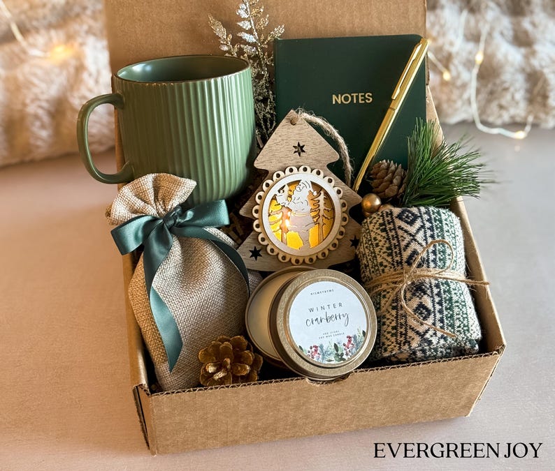 Puede incluir: Una caja de regalo llena con una taza verde, un cuaderno peque&ntilde;o con el texto "NOTES", un bol&iacute;grafo dorado, un adorno de &aacute;rbol de Navidad de madera, una vela, una bolsa peque&ntilde;a y una toalla estampada. La caja tiene el texto "EVERGREEN JOY" en la parte inferior.