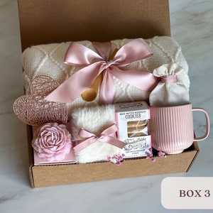May include: A gift box filled with self-care items, including a white blanket with a pink ribbon, a pink mug, a rose water meringue cookie package, a pink candle, a white faux fur pouch with a pink ribbon, and a small white bag. The box is labeled "BOX 3".
