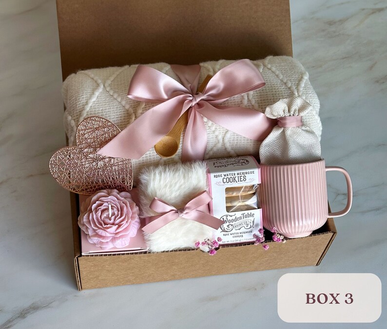 May include: A gift box filled with self-care items, including a white blanket with a pink ribbon, a pink mug, a rose water meringue cookie package, a pink candle, a white faux fur pouch with a pink ribbon, and a small white bag. The box is labeled "BOX 3".