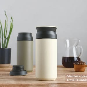 May include: Two stainless steel travel tumblers with black lids. One tumbler is beige and the other is green. The tumblers are on a wooden surface with coffee beans and a glass pitcher of coffee in the background.