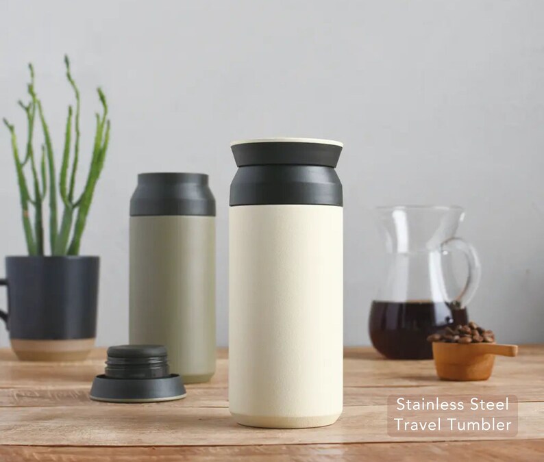 May include: Two stainless steel travel tumblers with black lids. One tumbler is beige and the other is green. The tumblers are on a wooden surface with coffee beans and a glass pitcher of coffee in the background.