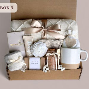 May include: A gift box labeled "Box 5" filled with self-care items, including a white blanket, a white mug, a bar of soap, a jar of bath salts, a white hand cream, a white flower, and a white and beige pouch with a heart.