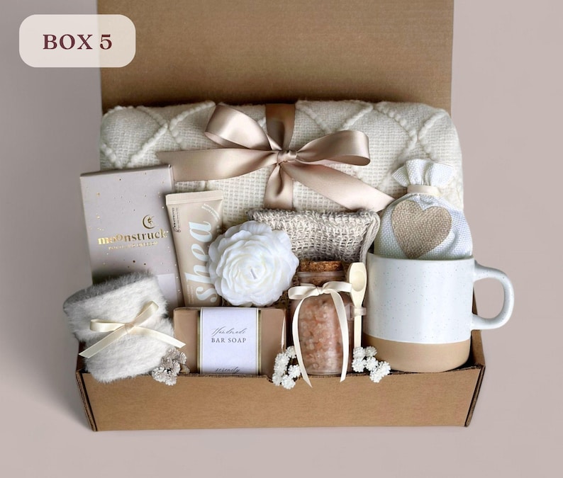 May include: A gift box labeled "Box 5" filled with self-care items, including a white blanket, a white mug, a bar of soap, a jar of bath salts, a white hand cream, a white flower, and a white and beige pouch with a heart.