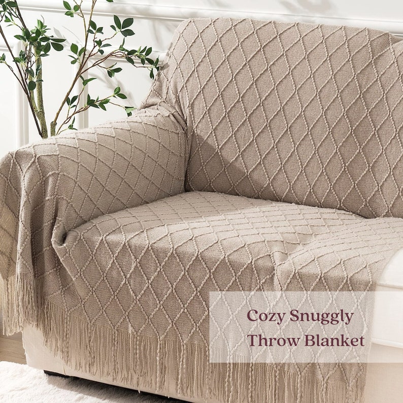 Puede incluir: Una manta beige con un patr&oacute;n de diamantes y flecos, colocada sobre un sof&aacute;. El texto "Cozy Snuggly Throw Blanket" est&aacute; en un recuadro blanco.