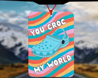 May include: A car air freshener with a rainbow background and a blue and white cartoon clog. The text "You Croc My World" is written on the air freshener.