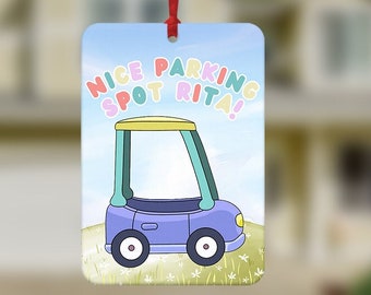 May include: A rectangular car air freshener with a cartoon car design. The car is purple with a yellow roof and green window frames, set against a blue sky and green grass background. Text reads "NICE PARKING SPOT RITA!"