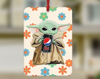 May include: A car air freshener featuring a green cartoon character holding a can of Pepsi with the text "Pepsi" on it. The character is on a white background with a pattern of orange and blue flowers.