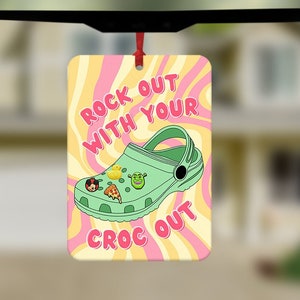 May include: A green and white cartoon-style clog with various charms attached. The background is pink and yellow with a swirl pattern. The text "Rock Out With Your Croc Out" is written in pink and white.