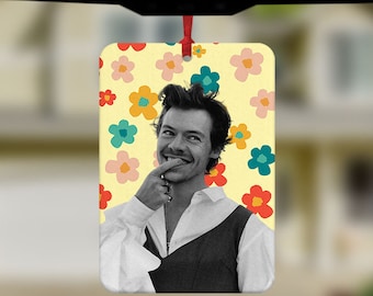 May include: A car air freshener featuring a black and white photo of a person with a light brown beard and a light blue shirt. The background is a yellow pattern of colorful flowers.