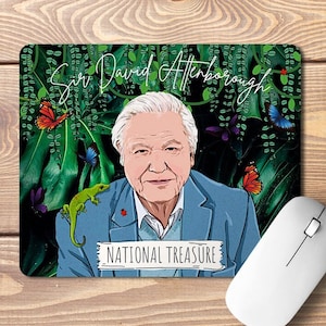 Op de afbeelding: Een muismat met een kleurrijke illustratie van Sir David Attenborough met een groene hagedis op zijn schouder. De achtergrond is een weelderige groene jungle met vlinders en wijnstokken. De tekst "National Treasure" staat onder Attenborough's afbeelding.