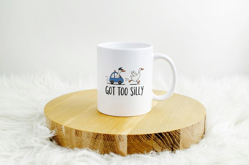 May include: White ceramic mug with a whimsical design. The mug features a cartoon goose being chased by a goose in a police car, with the text "GOT TOO SILLY" below. The mug is on a wooden round stand.