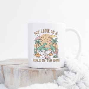 May include: White ceramic mug with a whimsical dinosaur design. The illustration features palm trees, a crescent moon, and the text "MY LIFE IS A WALK IN THE PARK". The design includes various colorful dinosaurs.