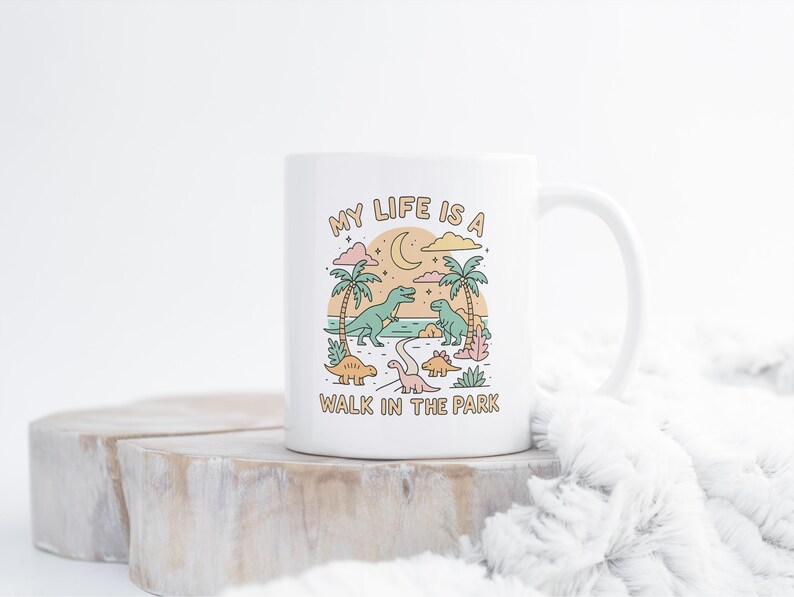 May include: White ceramic mug with a whimsical dinosaur design. The illustration features palm trees, a crescent moon, and the text "MY LIFE IS A WALK IN THE PARK". The design includes various colorful dinosaurs.