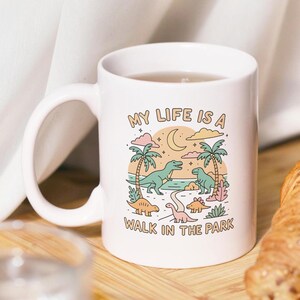 May include: White ceramic mug with a whimsical dinosaur illustration. The design features a beach scene with palm trees, dinosaurs, a crescent moon, and the text "MY LIFE IS A WALK IN THE PARK".