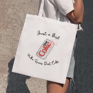 May include: White tote bag with a Diet Coke can illustration and a pink bow. The text reads "Just a Girl Who Loves Diet Coke" in black script. The bag is held by a person.
