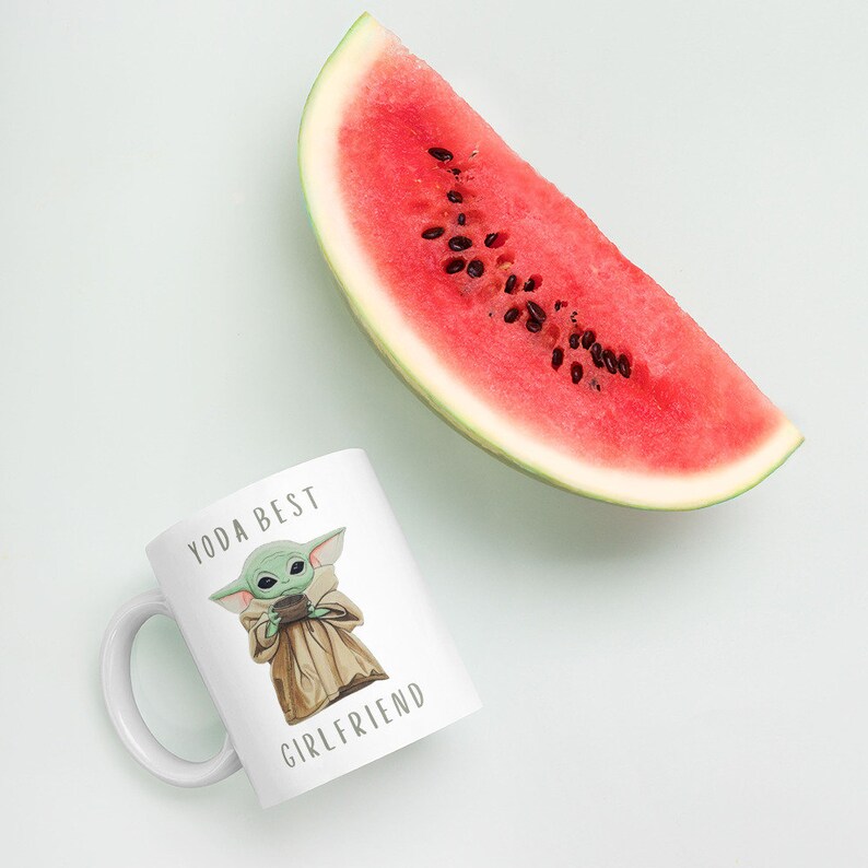 Op de afbeelding: Een witte keramische mok met een groen cartoonfiguur dat een kopje vasthoudt. Op de mok staat de tekst "Yoda Best Girlfriend". Op de achtergrond is een plak watermeloen te zien.