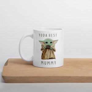May include: White ceramic mug with a green and brown cartoon character holding a cup. The text "YODA BEST MUMMY" is printed on the mug.