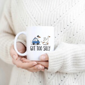 May include: White ceramic mug with a whimsical design featuring a goose driving a blue car and another goose running. The text "GOT TOO SILLY" is printed below the illustration. The mug is held by a person.