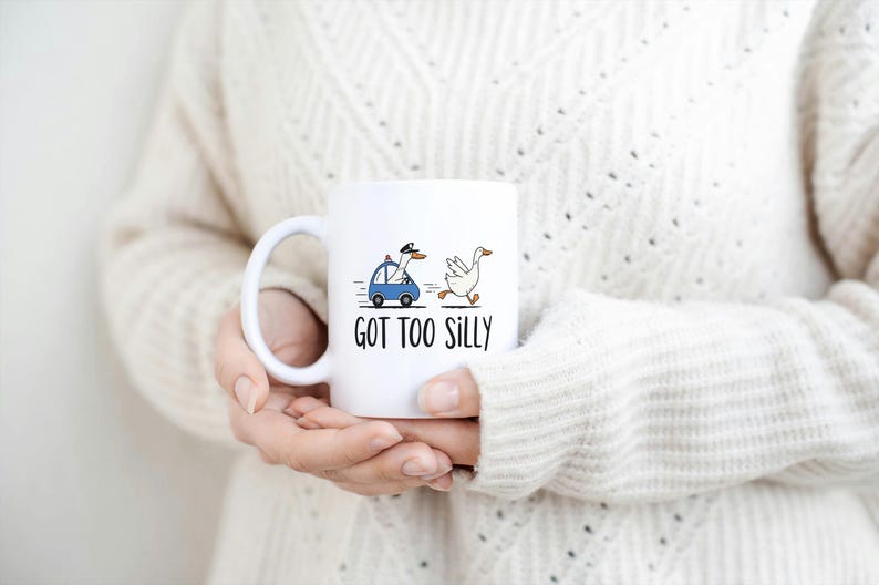 May include: White ceramic mug with a whimsical design featuring a goose driving a blue car and another goose running. The text "GOT TOO SILLY" is printed below the illustration. The mug is held by a person.