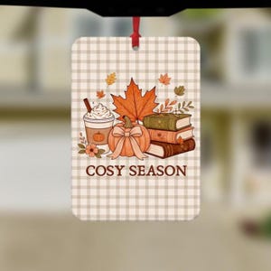 May include: A rectangular hanging air freshener featuring a beige and white checkered background. The design includes a coffee cup, a pumpkin with a bow, a stack of three books, and autumn leaves. The text COSY SEASON is printed in brown.