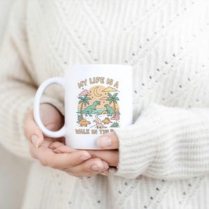 May include: White ceramic mug with a handle featuring a colorful illustration of dinosaurs, palm trees, and a crescent moon. The text "MY LIFE IS A WALK IN THE PARK" is printed on the mug. The mug is held by a person wearing a cream-colored sweater.
