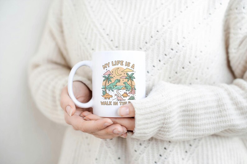 May include: White ceramic mug with a handle featuring a colorful illustration of dinosaurs, palm trees, and a crescent moon. The text "MY LIFE IS A WALK IN THE PARK" is printed on the mug. The mug is held by a person wearing a cream-colored sweater.