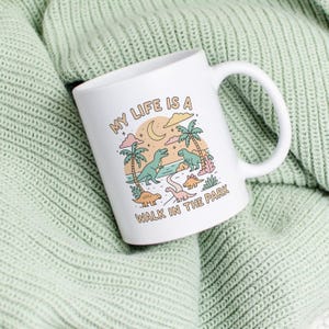 May include: White ceramic mug with a wraparound illustration of dinosaurs, palm trees, and a crescent moon. The text "MY LIFE IS A WALK IN THE PARK" is printed on the mug. The mug has a curved handle and is set against a light green knitted background.
