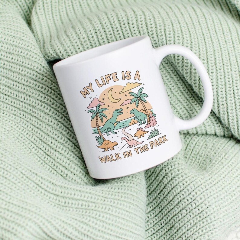 May include: White ceramic mug with a wraparound illustration of dinosaurs, palm trees, and a crescent moon. The text "MY LIFE IS A WALK IN THE PARK" is printed on the mug. The mug has a curved handle and is set against a light green knitted background.