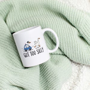 May include: White ceramic mug with a whimsical illustration of two geese, one driving a blue car and the other running. The text "GOT TOO SILLY" is printed below the geese. The mug is set on a light green knitted blanket.