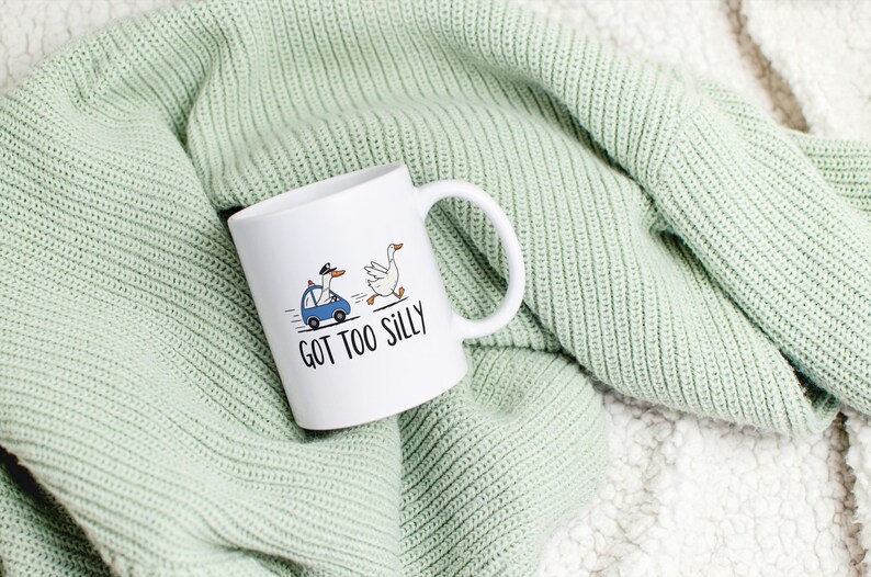 May include: White ceramic mug with a whimsical illustration of two geese, one driving a blue car and the other running. The text "GOT TOO SILLY" is printed below the geese. The mug is set on a light green knitted blanket.