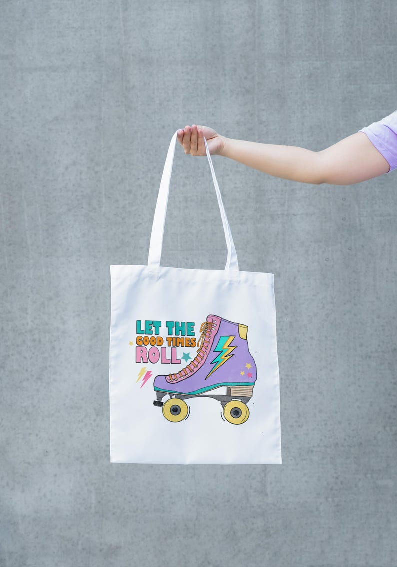 May include: White tote bag with a purple roller skate design and the text "LET THE GOOD TIMES ROLL". The roller skate has yellow wheels. The bag is held up by a person.