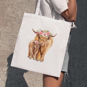 May include: White tote bag featuring a watercolor illustration of a Highland cow and calf, both adorned with floral crowns. The cows have long, shaggy brown fur. The bag is held by a person.