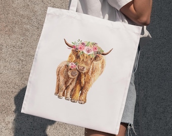 May include: White tote bag featuring a watercolor illustration of a Highland cow and calf, both adorned with floral crowns. The cows have long, shaggy brown fur. The bag is held by a person.
