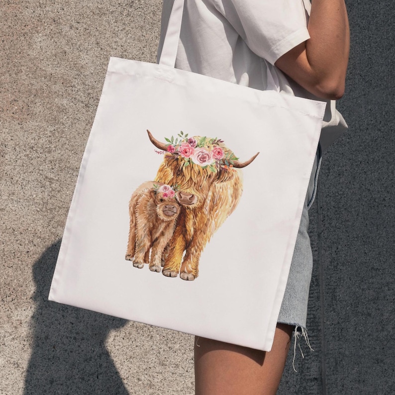 May include: White tote bag featuring a watercolor illustration of a Highland cow and calf, both adorned with floral crowns. The cows have long, shaggy brown fur. The bag is held by a person.