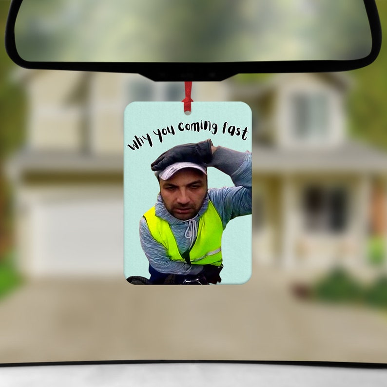 K&ouml;nnte beinhalten: Rechteckiger Auto-Lufterfrischer mit einem Foto eines Mannes in einer Sicherheitsweste und Kappe, mit dem Text "why you coming fast". Der Hintergrund ist hellblau.