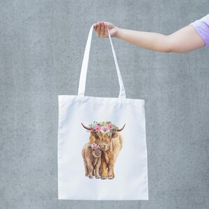May include: White tote bag featuring a watercolor illustration of a Highland cow and calf, both adorned with floral crowns. The bag has long white handles and is held up against a gray background.