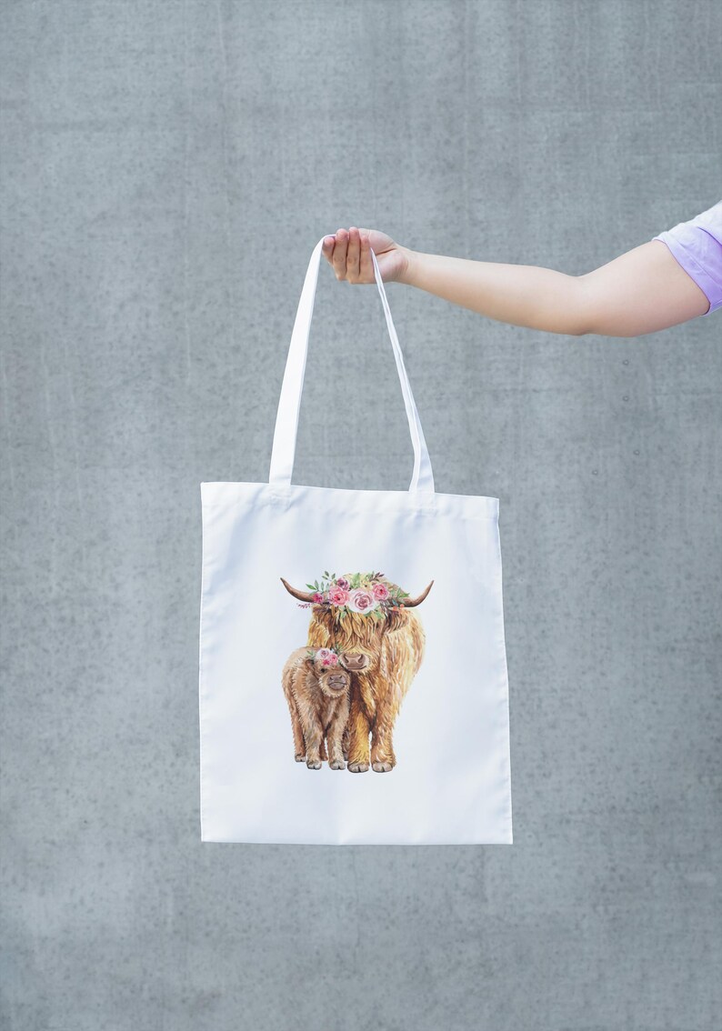 May include: White tote bag featuring a watercolor illustration of a Highland cow and calf, both adorned with floral crowns. The bag has long white handles and is held up against a gray background.