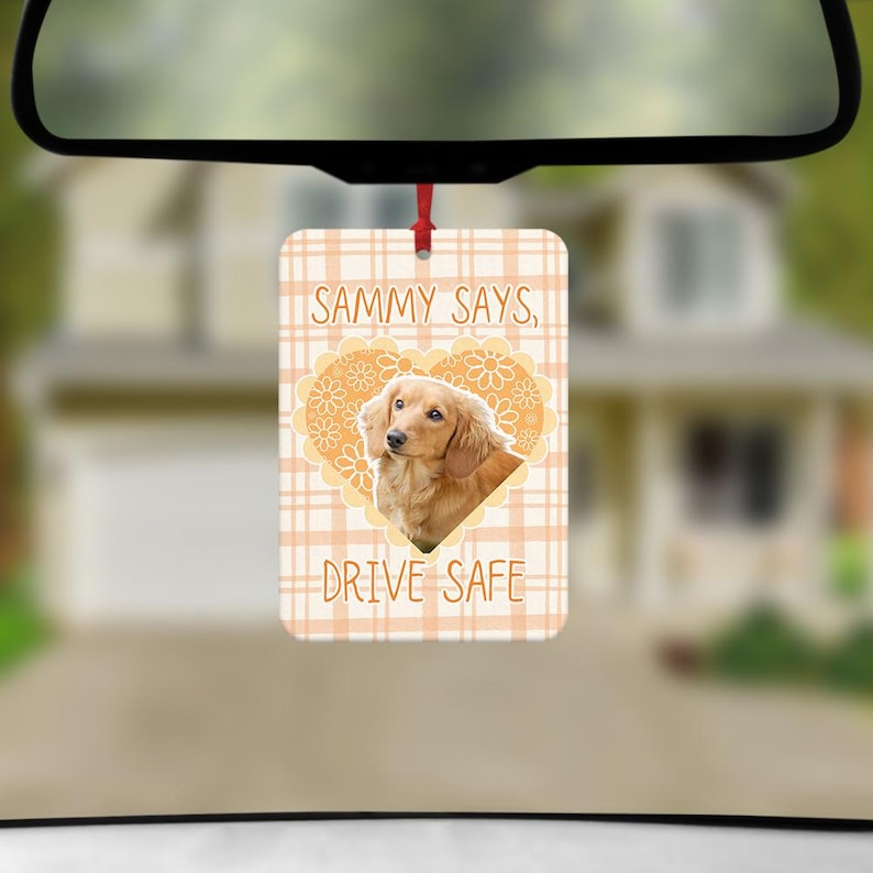 May include: A rectangular car air freshener featuring a light orange plaid background. Centered is a heart-shaped floral frame containing a photograph of a long-haired light brown dog. The text reads SAMMY SAYS, DRIVE SAFE in brown lettering.