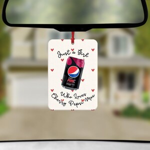 May include: A rectangular car air freshener hanging from a red string, featuring a white background with small red hearts. In the center is a black Pepsi Max Cherry soda can graphic with the text Just a Girl Who Loves Cherry Pepsi Max.