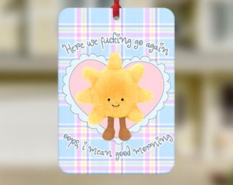 May include: A rectangular ornament with a blue plaid background and a plush yellow sun with brown legs. The ornament has the text "Here we fucking go again" and "oops, I mean good morning". A red ribbon is attached for hanging.