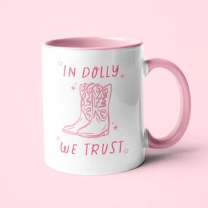 May include: A white ceramic mug with a pink interior and handle. The front features a pink line-art illustration of two cowboy boots surrounded by small stars. The text "IN DOLLY WE TRUST" is printed in pink lettering above and below the boots.