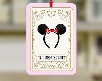 May include: A rectangular hanging air freshener with a light pink border. The center features a cream-colored tarot card design with gold filigree, a black mouse ear headband with a red and white polka dot bow, and the text THE DISNEY ADULT.
