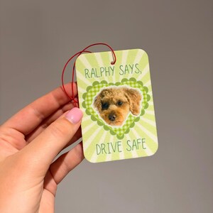 May include: A rectangular air freshener with a light green and white sunburst background. It features a scalloped green heart frame containing a photograph of a brown dog. The text reads RALPHY SAYS, DRIVE SAFE in green capital letters.