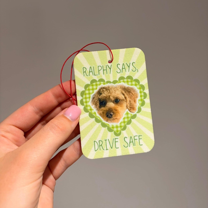 May include: A rectangular air freshener with a light green and white sunburst background. It features a scalloped green heart frame containing a photograph of a brown dog. The text reads RALPHY SAYS, DRIVE SAFE in green capital letters.
