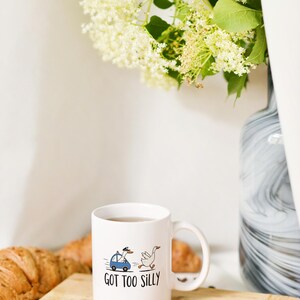 May include: White ceramic mug with a whimsical illustration of a goose being chased by a police car with a goose driver. The mug has the text "GOT TOO SILLY". A croissant, spoon, and coffee beans are on a wooden board.