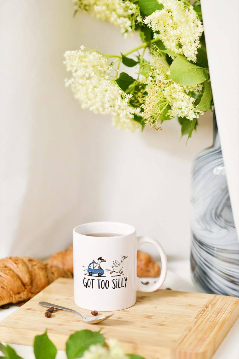 May include: White ceramic mug with a whimsical illustration of a goose being chased by a police car with a goose driver. The mug has the text "GOT TOO SILLY". A croissant, spoon, and coffee beans are on a wooden board.