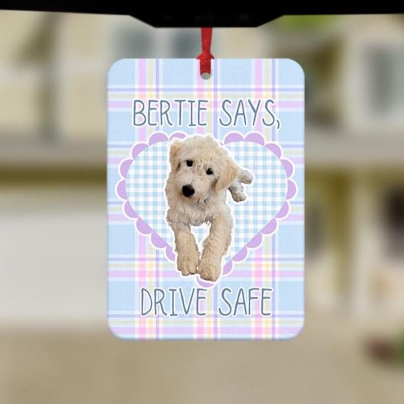 May include: A rectangular hanging air freshener featuring a light blue, white, and yellow plaid background. A central lavender heart frame surrounds a photo of a cream-colored doodle dog. The text reads, BERTIE SAYS, DRIVE SAFE, in blue.