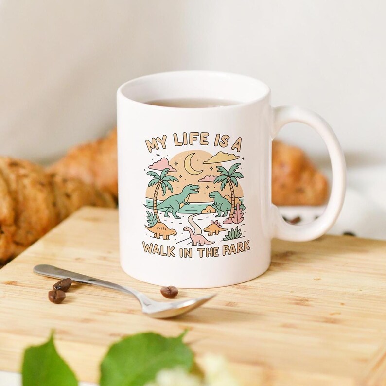 May include: White ceramic mug with a wraparound illustration featuring dinosaurs, palm trees, and the text "MY LIFE IS A WALK IN THE PARK." The design includes a crescent moon and clouds. A spoon and coffee beans are in the foreground.